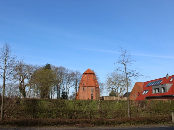 The ruins of a windmill in Haßbergen, surrounded by trees and modern houses under a blue sky.