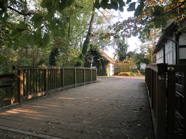 Brücke an der Bunten Mühle Holzbrücke im Grünen, umgeben von Bäumen und Laub, führt zu weißen Fachwerkhäusern im Hintergrund.