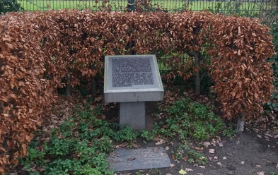 Gedenktafel Steinplatte mit Gravur, umgeben von herbstlich verfärbten Buchenhecken, steht auf grasbewachsenem Boden.