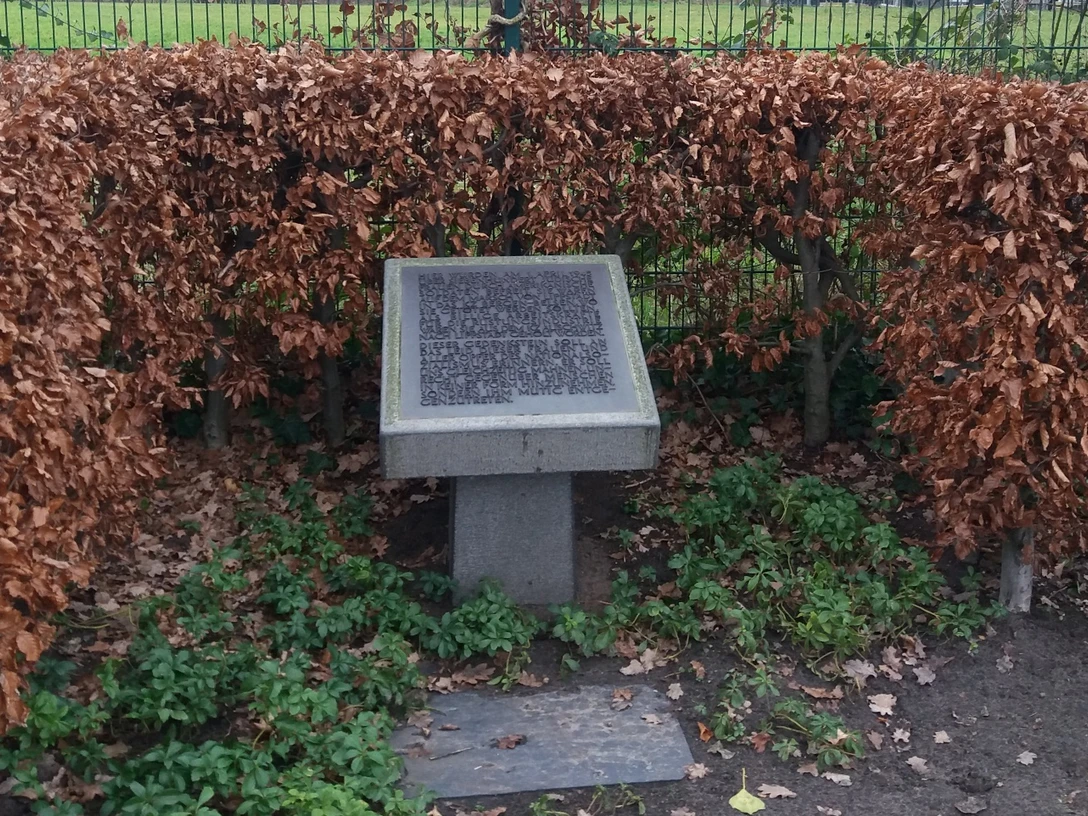 Gedenktafel Steinplatte mit Gravur, umgeben von herbstlich verfärbten Buchenhecken, steht auf grasbewachsenem Boden.
