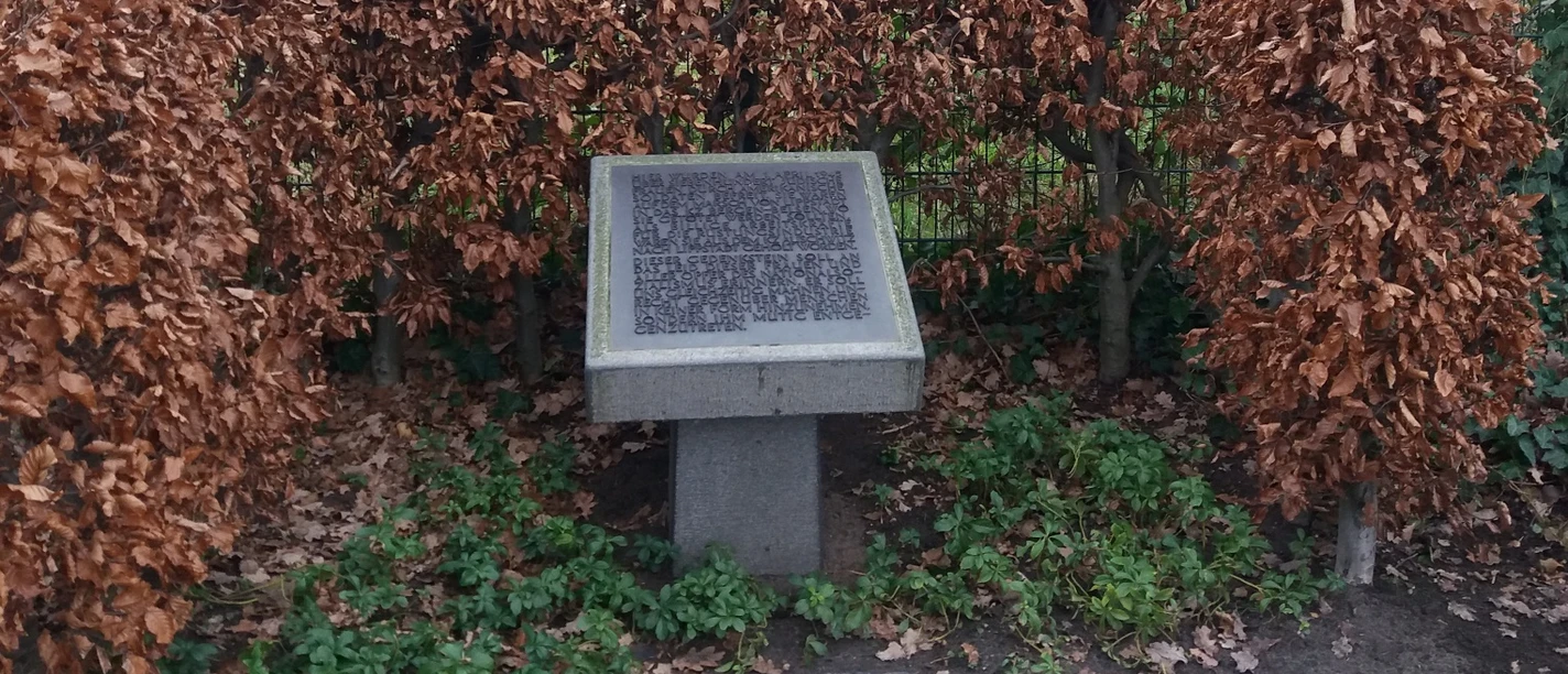 Gedenktafel Steinplatte mit Gravur, umgeben von herbstlich verfärbten Buchenhecken, steht auf grasbewachsenem Boden.