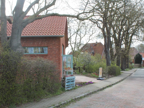 Aan een geplaveide straat staat een gebouw van rode baksteen met een rood pannendak. Voor het gebouw ligt een smal voetpad met bomen en struiken. Op de voorgrond staat een telefooncel.