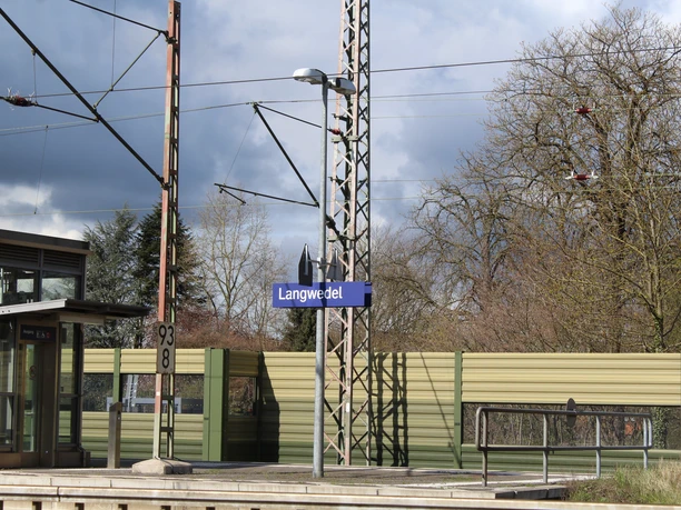 Bahnhof Langwedel Bahnhof Langwedel mit Bahnsteig, Signalmast, Zuggleis und grünen Lärmschutzwänden unter wolkigem Himmel.