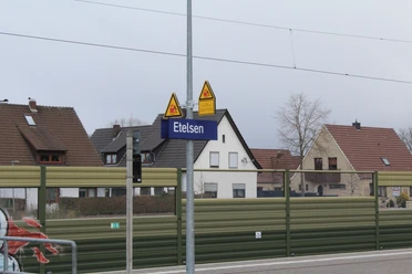 Bahnhofsschild "Etelsen" vor Wohngebäuden, im Hintergrund teils bewölkter Himmel und Oberleitungen.