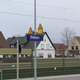 Bahnhofsschild "Etelsen" vor Wohngebäuden, im Hintergrund teils bewölkter Himmel und Oberleitungen.