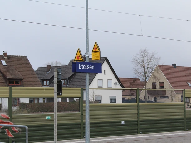 Bahnhof Etelsen Bahnhofsschild "Etelsen" vor Wohngebäuden, im Hintergrund teils bewölkter Himmel und Oberleitungen.