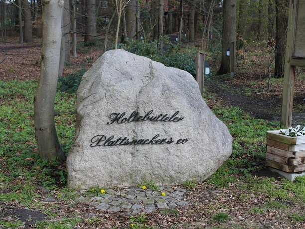 A large natural stone with the black inscription "Holtebütteler Plattsnacker ev" stands in the park.