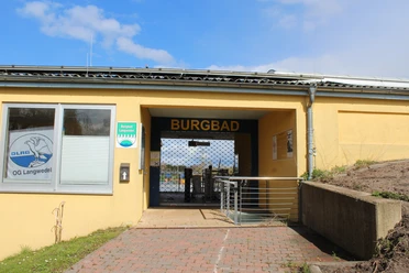Eingangsbereich des Burgbads Langwedel mit gelbem Gebäude, Barrierefreiheitshinweis und Tor.