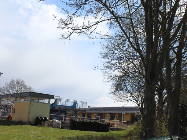 An outdoor pool in a green setting with slides, a changing room building, and trees in the spring.