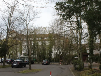 Kurklinik Bad Hopfenberg Großer Parkplatz mit Autos vor einem dreistöckigen Gebäude, umgeben von kahlen Bäumen im Frühjahr.