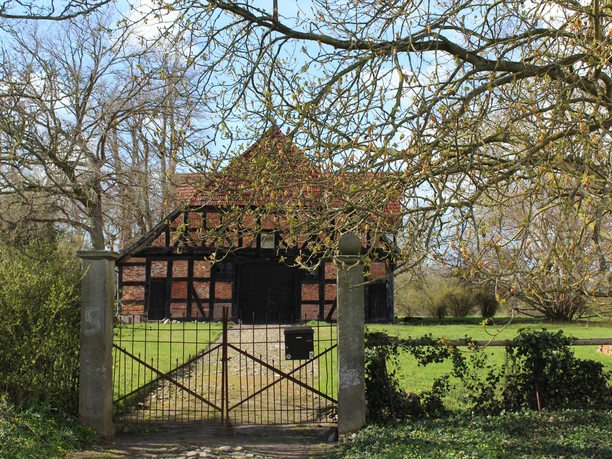 Fachwerkhaus mit Backsteinmauern hinter einem schmiedeeisernen Tor, umgeben von grüner Landschaft.