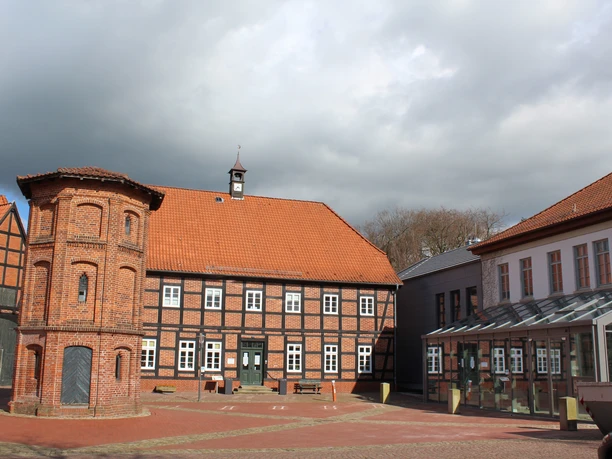 Rotes Backsteingebäude mit Fachwerk, rotem Ziegeldach und Wendelturm bei bewölktem Himmel.