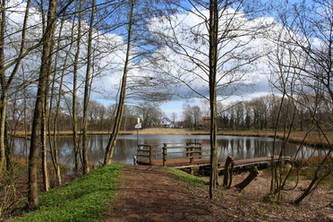 Böttchers Moor Ein ruhiger Teich im Böttchers Moor mit Holzstegen, umgeben von kargen Bäumen unter blauem Himmel.