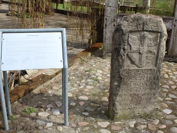 Steinmonument mit reliefierter Wappeninschrift, umgeben von Kopfsteinpflaster und Informationstafel.
