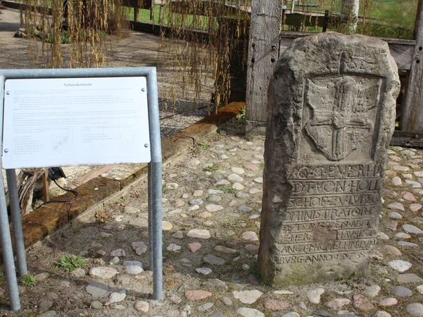 Steinmonument mit reliefierter Wappeninschrift, umgeben von Kopfsteinpflaster und Informationstafel.