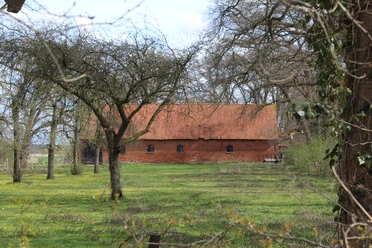 Gut Donnerhorst Ein Backsteinhaus mit rotem Dach steht hinter Bäumen auf einer grünen Wiese im Frühling.