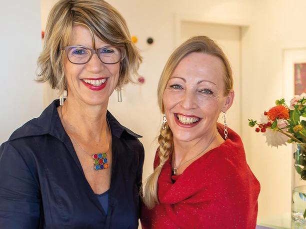 Sigrid-Carmen-hotel-eikamper-hoehe.jpg Zwei lächelnde Frauen in eleganter Kleidung präsentieren jeweils einen Apfel in freundlicher Atmosphäre.