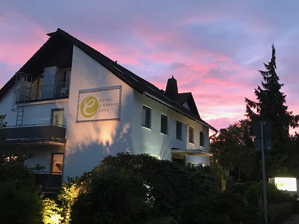 Hotelansicht Fassade eines Hotels bei Sonnenuntergang, beleuchtet mit sanften Lichtern, im Vordergrund Grünflächen.