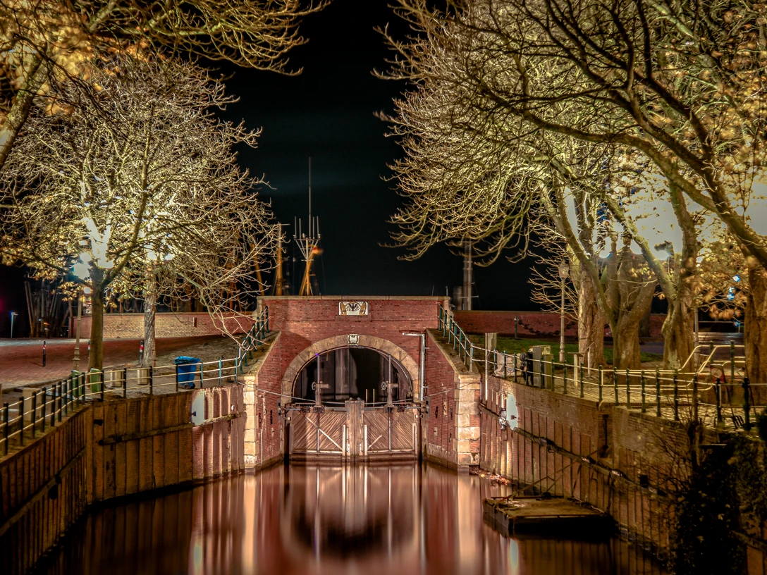 Siel in Greetsiel Historisches Sieltor in Greetsiel bei Nacht, beleuchtet von warmem Licht, umgeben von kahlen Bäumen.