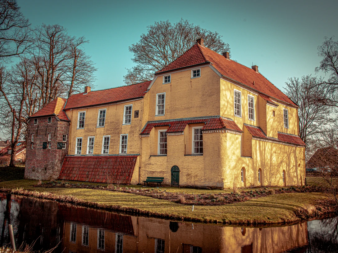 Manningaburg in Pewsum Backsteinbau einer Burg mit gelbem Putz und rotem Dach, von Wassergraben und kahlen Bäumen umgeben.