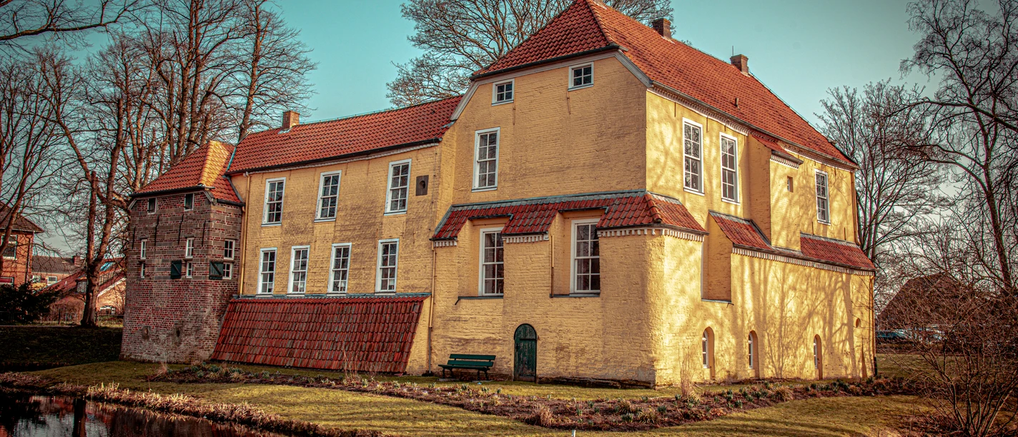 Manningaburg in Pewsum Backsteinbau einer Burg mit gelbem Putz und rotem Dach, von Wassergraben und kahlen Bäumen umgeben.