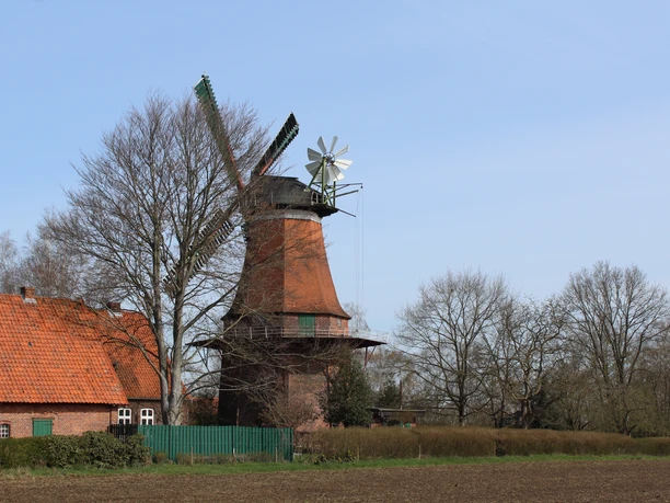 A windmill with a red brick building and green blades, surrounded by trees and vast fields.