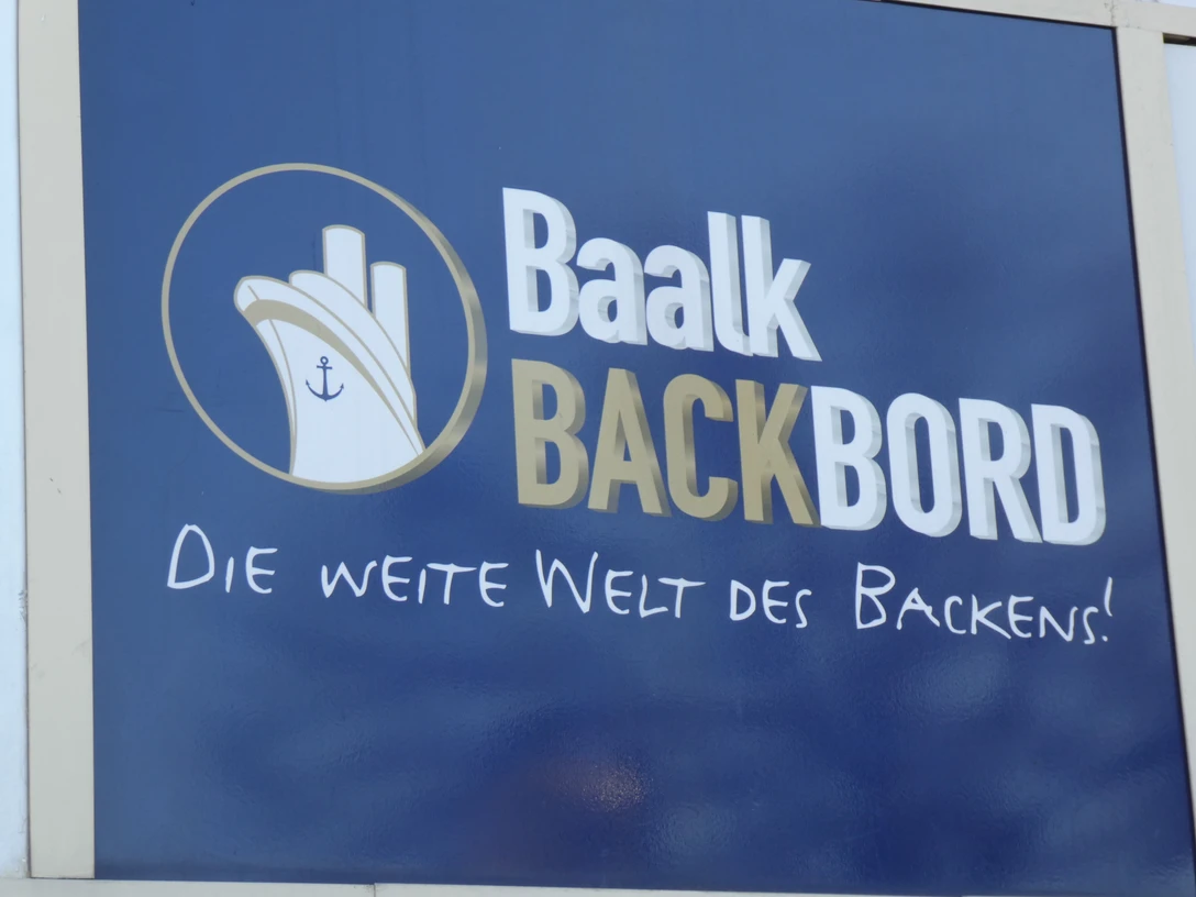 Bäckerei Baalk Baalk Backbord Logo auf blauem Hintergrund mit dem Slogan "Die weite Welt des Backens!".