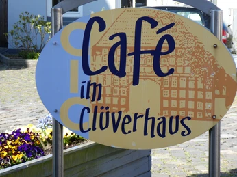 Schild des Cafés im Clüverhaus inmitten eines gepflasterten Hofes, umgeben von Blumen.