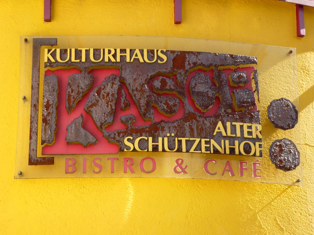 Rostiges Metallschild mit der Aufschrift "Kulturhaus KASCH Alter Schützenhof Bistro & Café" an gelber Wand.