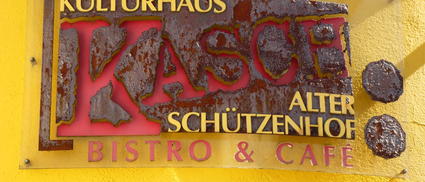 Gastro im KASCH Rostiges Metallschild mit der Aufschrift "Kulturhaus KASCH Alter Schützenhof Bistro & Café" an gelber Wand.