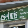 Les Amis Grünes Schild von "Les Amis" mit den Angeboten Pizza, Baguetterie und Salate, auf Backsteinwand.