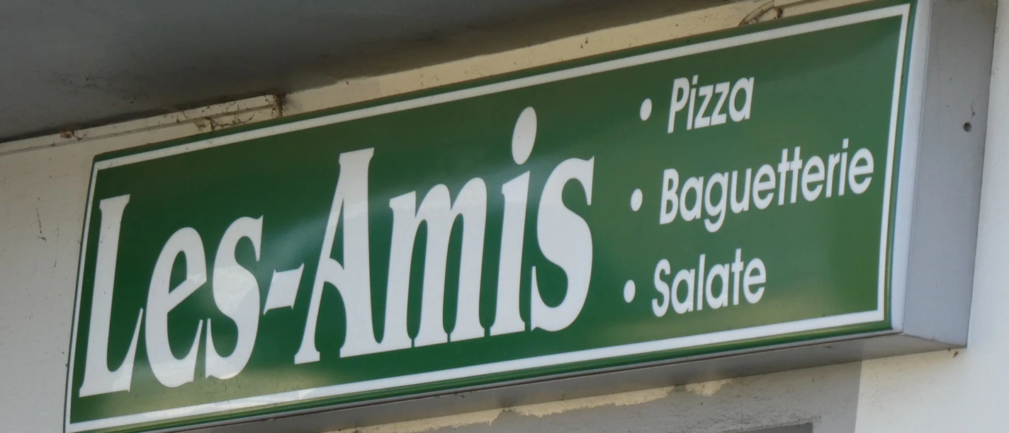 Les Amis Grünes Schild von "Les Amis" mit den Angeboten Pizza, Baguetterie und Salate, auf Backsteinwand.