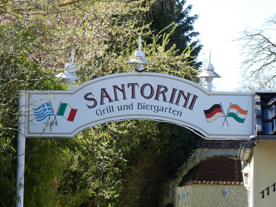 Das Eingangsschild des Restaurants Santorini zeigt eine griechische und deutsche Flagge mit grüner Umgebung.The entrance sign of the Santorini restaurant shows a Greek and German flag with green surroundings.Indgangsskiltet til Santorini-restauranten viser et græsk og et tysk flag med grønne omgivelser.Het toegangsbord van het Santorini-restaurant toont een Griekse en Duitse vlag met een groene achtergrond.