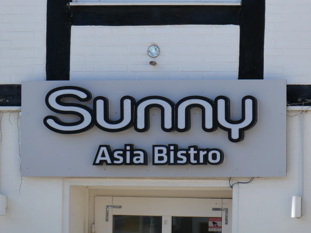 Leuchtendes Schild mit der Aufschrift "Sunny Asia Bistro" an einer Gebäudefassade mit Fachwerkelementen.Illuminated sign with the inscription "Sunny Asia Bistro" on a building façade with half-timbered elements.Belyst skilt med påskriften "Sunny Asia Bistro" på en bygningsfacade med bindingsværkselementer.Verlicht uithangbord met het opschrift "Sunny Asia Bistro" op een gevel met vakwerkelementen.
