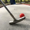 Ein silberner Minigolfschläger im Holzstil trifft auf einen roten Ball auf einer Betonanlage.
