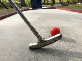 Minigolf Ein silberner Minigolfschläger im Holzstil trifft auf einen roten Ball auf einer Betonanlage.