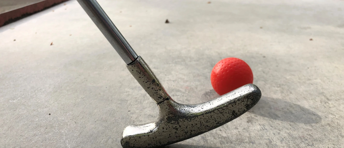 Minigolf Ein silberner Minigolfschläger im Holzstil trifft auf einen roten Ball auf einer Betonanlage.