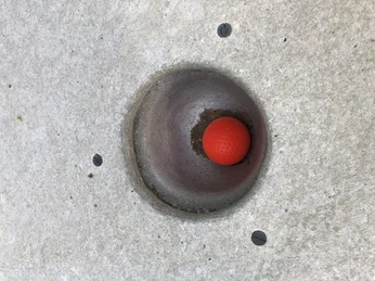 Roter Golfball liegt in einem Betonloch auf einer Minigolfanlage, umgeben von einer glatten Oberfläche.
