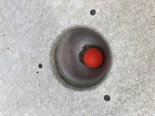Roter Golfball liegt in einem Betonloch auf einer Minigolfanlage, umgeben von einer glatten Oberfläche.