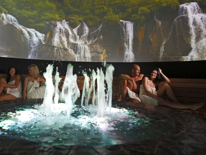H2O Sport- und Freizeitbad Herford-Vesitorni Sauna Menschen entspannen in einer Sauna mit Wasserfontäne, umgeben von einem Wandbild mit Wasserfällen.