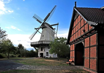 TonnenheiderHochzeitsmühle Historische Windmühle mit drehenden Flügeln, umgeben von Bäumen, neben einem roten Fachwerkgebäude.