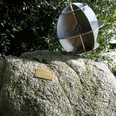 Metallskulptur auf großem Stein, umgeben von Grün, mit kleinem Messingschild auf sonniger Wiese.
