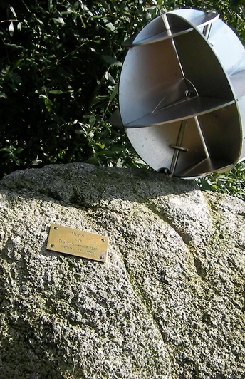 Metallskulptur auf großem Stein, umgeben von Grün, mit kleinem Messingschild auf sonniger Wiese.