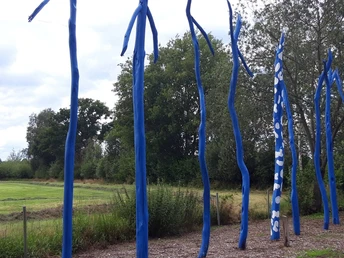 Blaue Skulpturen ragen vor einem bewaldeten Hintergrund in den Himmel und säumen einen Wiesenweg.
