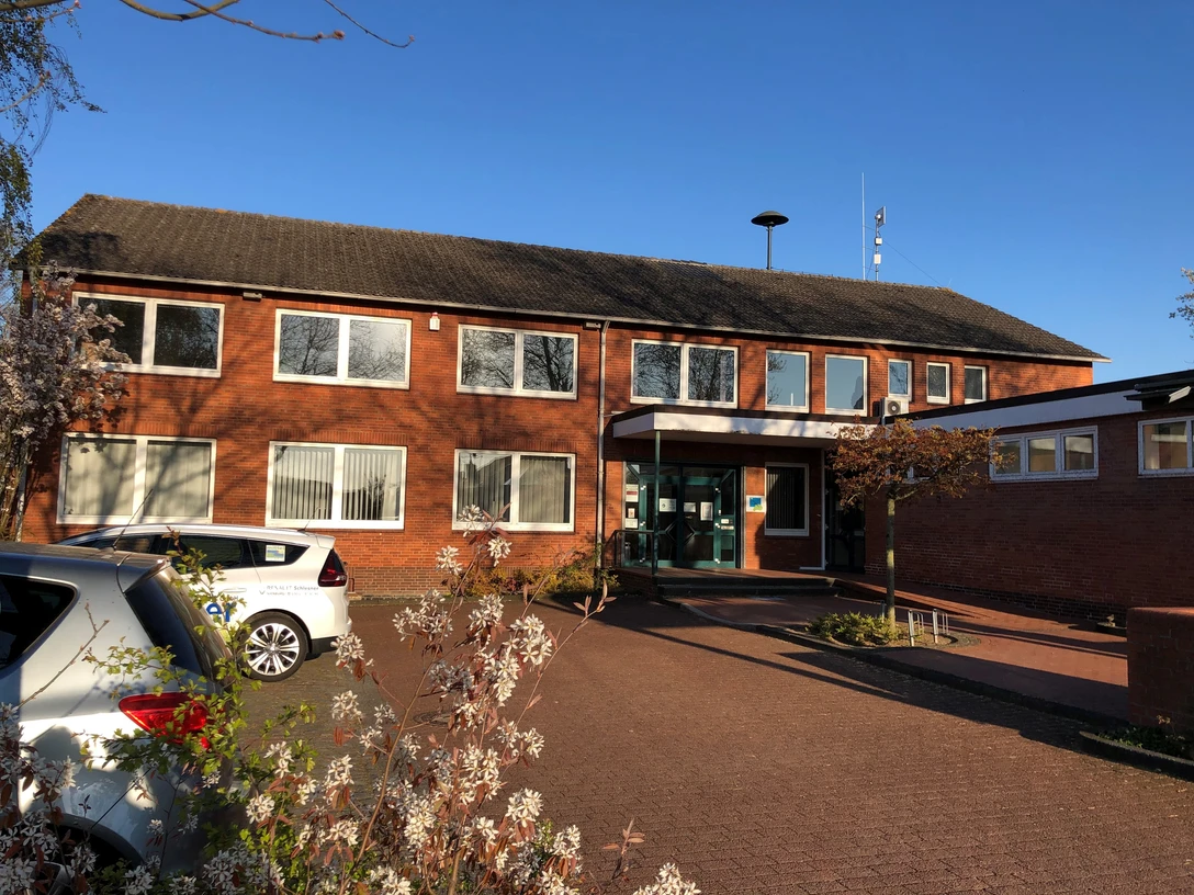 Backsteingebäude mit zwei Etagen, mehreren Fenstern und Eingangstür, umgeben von Büschen und Autos.