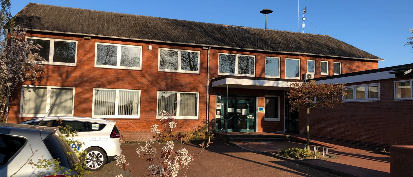 Backsteingebäude mit zwei Etagen, mehreren Fenstern und Eingangstür, umgeben von Büschen und Autos.