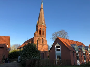 Rote Backsteinkirche in Estorf mit hohem Turm, umgeben von Gebäuden und blühenden Sträuchern.
