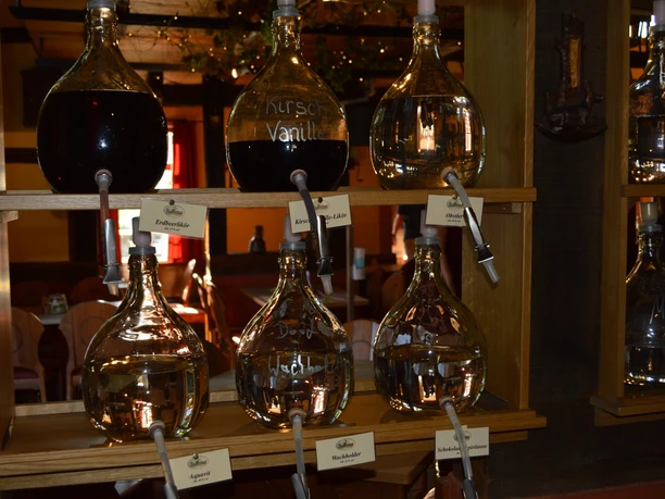 Liebharts Fachwerkdorf-Sudhaus 1 Verschiedene Glasballons mit Spirituosen und Schnaps, etikettiert und in einem rustikalen Regal arrangiert.