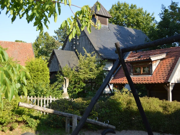 Liebharts Fachwerkdorf-Dorfkapelle Hinteransicht Rückansicht einer rauchgrauen Fachwerkkapelle umgeben von Grün und angrenzenden Fachwerkgebäuden.