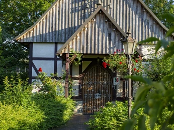 Liebharts Fachwerkdorf-Dorfkapelle Dorfkapelle mit Fachwerk und Schindeldach, umgeben von grünen Sträuchern und blühenden Rosen.
